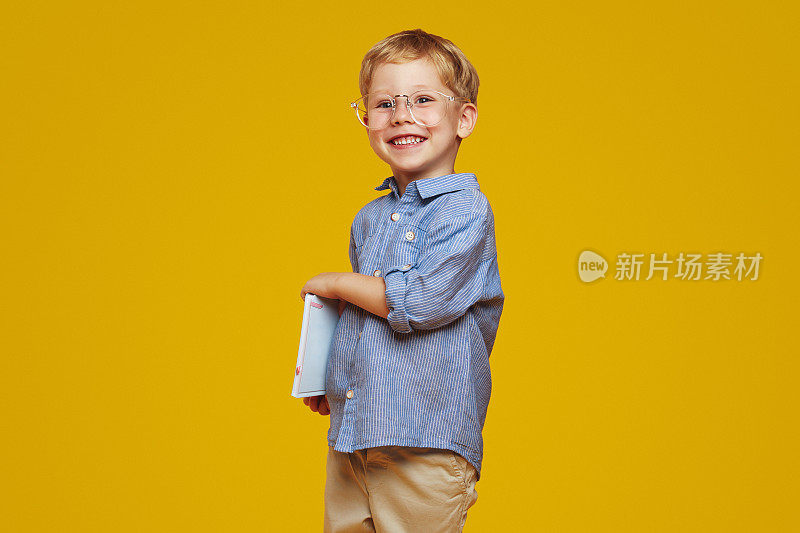 聪明的小男生戴着书呆子眼镜，穿着蓝色衬衫，拿着笔记本，对着镜头微笑