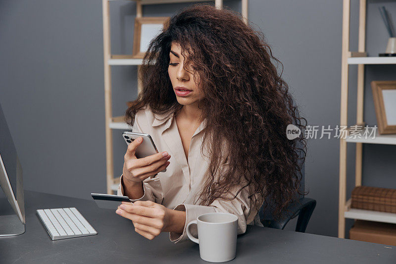 集中专注的棕褐色可爱卷曲的拉丁妇女亚麻衬衫小心地输入信用卡的详细信息在手机上。复制空间横幅。购物狂女士使用现代电脑进行网上购物。