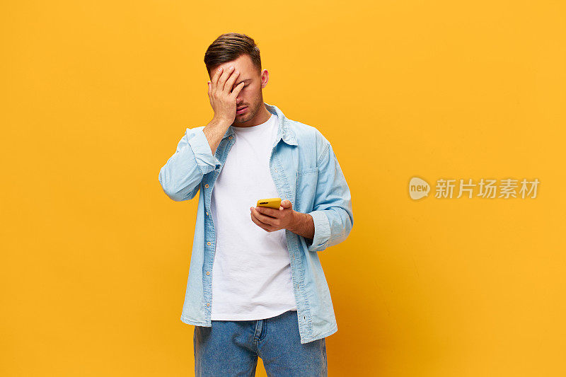沮丧，不高兴，晒黑英俊的男人在蓝色基本t恤得到坏的财务报告坏消息拿着手机摆出孤立的黄色工作室背景。复制空间横幅模型。在线社交媒体概念