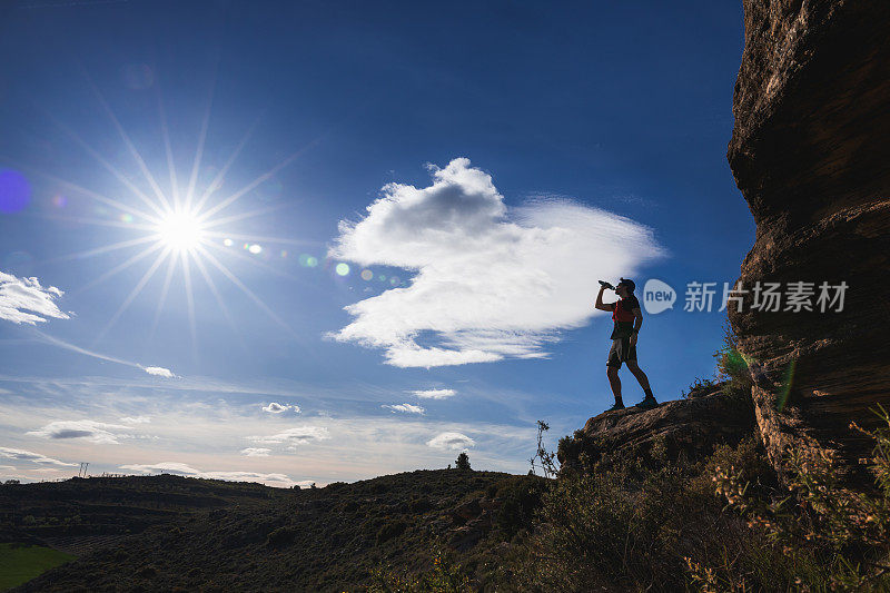 一名越野跑运动员站在一块崎岖的岩石上，拿着一瓶酒，周围是星形的太阳和蓝天白云的活力之美。