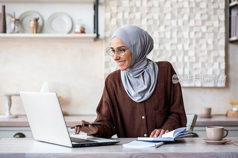 阿拉伯年轻女学生在家远程学习。他坐在餐桌旁的笔记本电脑前，在笔记本上写下总结
