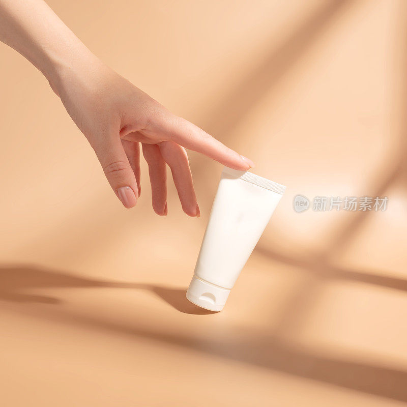 女人的手摸着一筒白色奶油的模型。未标示的化妆品包装在阳光下的米色背景。护肤概念。强烈的阴影