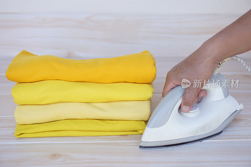 桌子上叠着一叠黄色衣服，手里拿着电熨斗。概念，日常琐事，家庭。熨烫和折叠衣服，使其整洁干净。维护和保管服装。