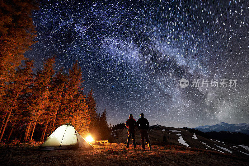 两名男性游客晚上在森林附近的露营中休息。小伙子们站在篝火和帐篷旁，在满是星星和银河的美丽夜空下，欣赏夜景。长时间曝光