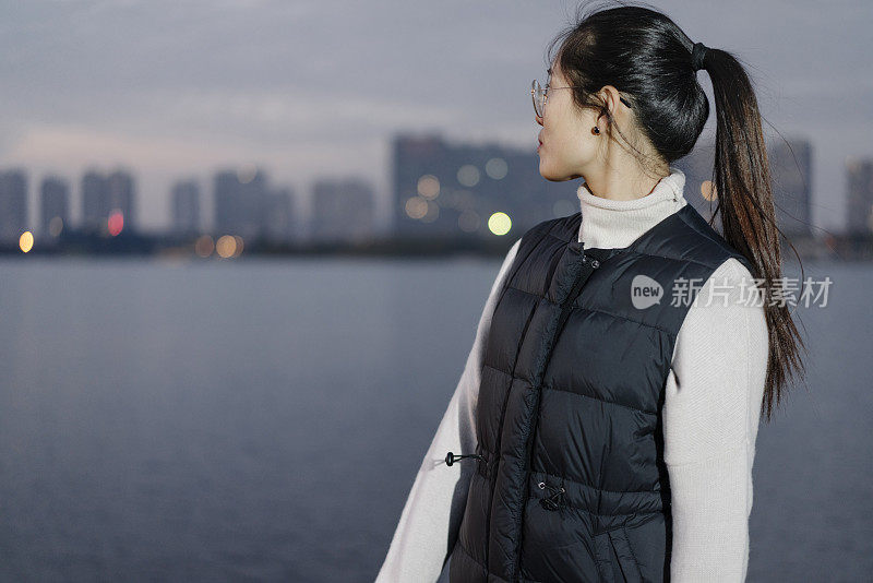 黄昏时分，一个女人站在湖边，背对着现代化的建筑物