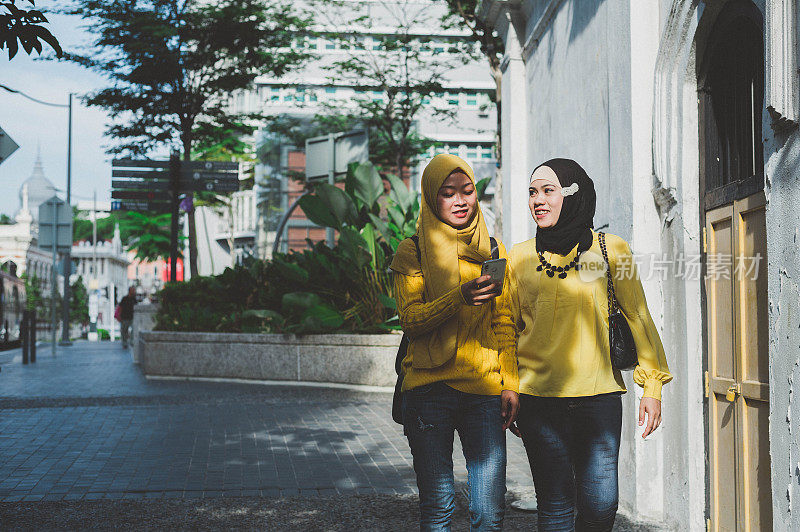 两名年轻的穆斯林妇女戴着头巾在城市街道上使用智能手机