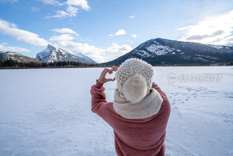 一名年轻女子在雪山松林上制作心形手指架，享受寒假的乐趣