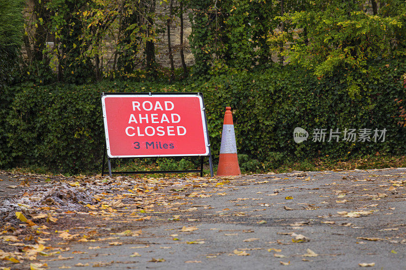 在科茨沃尔德的一条安静的乡村道路上，前面的道路关闭了警告标志，前景是秋叶。