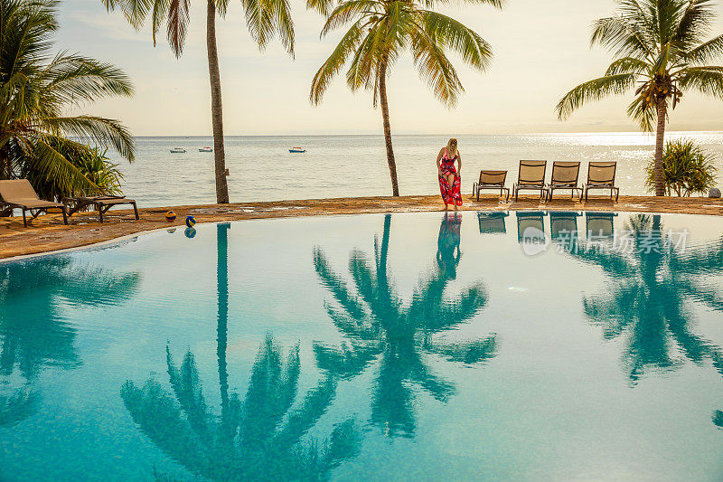 日落时分，坦桑尼亚桑给巴尔，一名身穿红色连衣裙的女子在棕榈树下的游泳池边散步