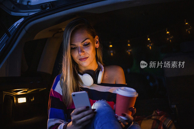一个年轻漂亮的女人露营在车的后面，放松，同时使用她的手机。