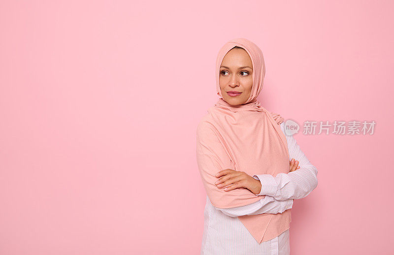 自信的阿拉伯穆斯林美丽女人的肖像，迷人的凝视和覆盖着粉红色头巾的头，站在四分之三的彩色背景与副本空间，并望向副本空间