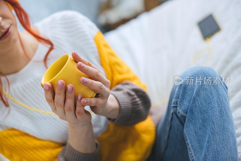 一个女孩坐在床上，喝着咖啡，戴着黄色耳机听着音乐。