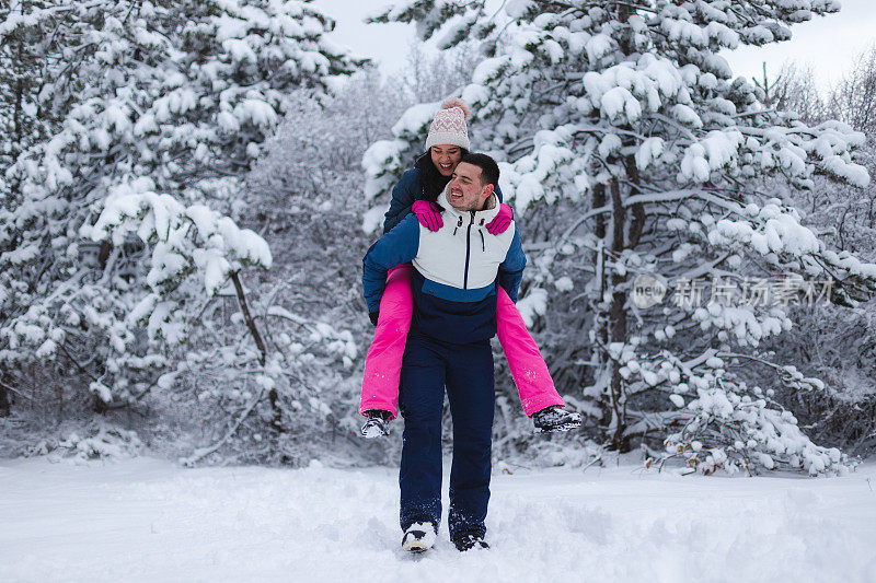 年轻的男朋友背着他的女朋友在雪地里玩，周围是雪树