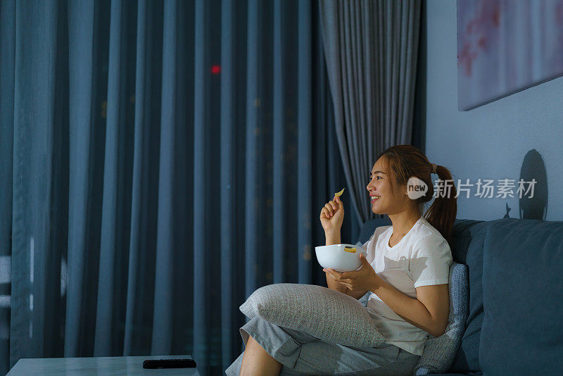 亚洲妇女看电视悬疑电影或新闻看起来快乐和有趣的和吃爆米花在家庭隔离时间在家里客厅沙发深夜。