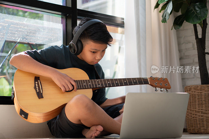 亚洲男孩一边玩吉他，一边在笔记本电脑上观看在线课程，同时在家练习在线学习音乐或乐器。男生通过视频通话在线学习，老师弹吉他。