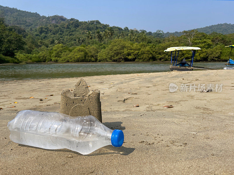 干地上用水桶和铁锹制作的沙堡外的塑料水瓶特写，沙滩，海岸上的海洋垃圾和污染，乱扔的沙子，渔船，有棕榈树的林地，重点在前景