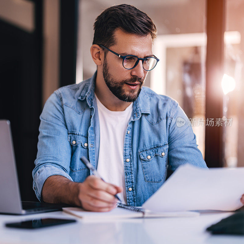 一个戴着眼镜的满意的年轻人坐在办公桌前，在他的工作场所做文书工作。