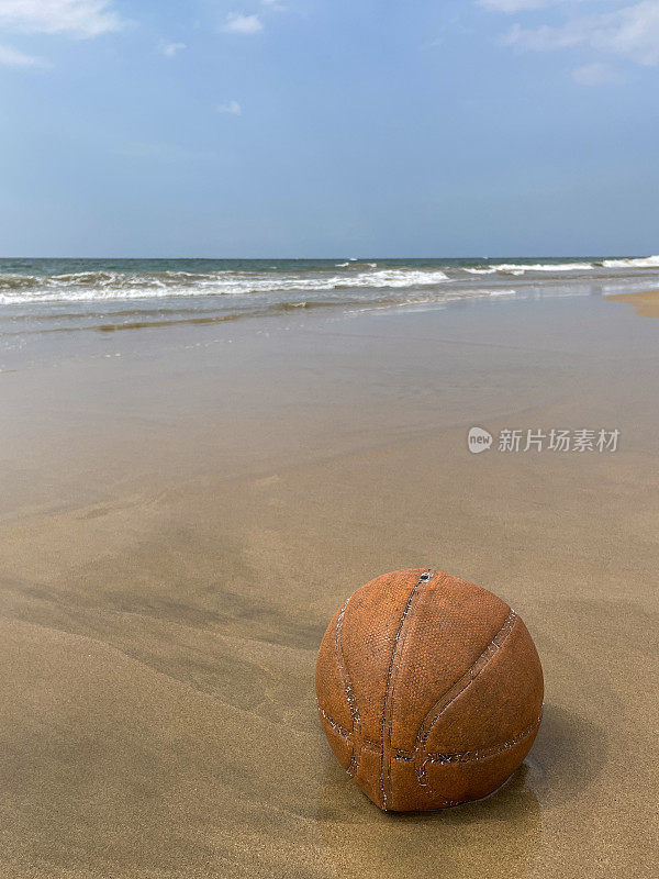 近距离图像，旧的，泄气的篮球被冲到沙滩上在退潮水的边缘，破碎的海浪冲刷海洋垃圾和岸上的污染，乱扔的沙子，肮脏的海滩，关注前景，复制空间
