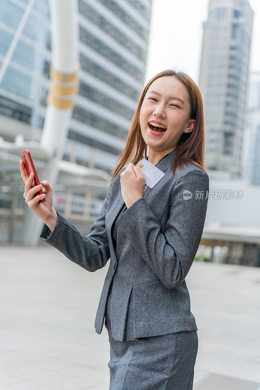 一名年轻的亚洲女商人穿着灰色的办公套装，在商业区用她的智能手机使用信用卡进行在线交易，她微笑着自信