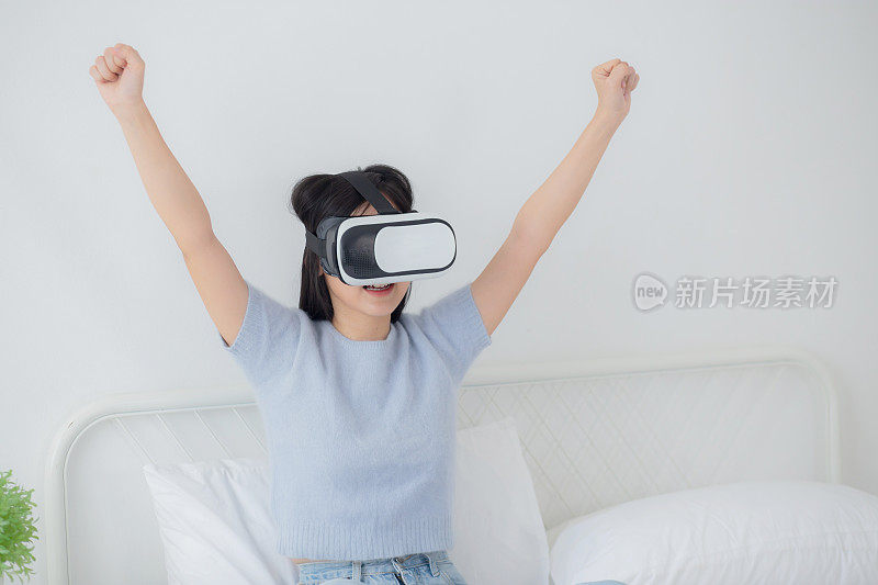 年轻的亚洲女性戴着vr耳机在家里的卧室里兴奋地玩游戏，女性使用虚拟现实或元宇宙创新来模拟3D，生活方式和技术概念。