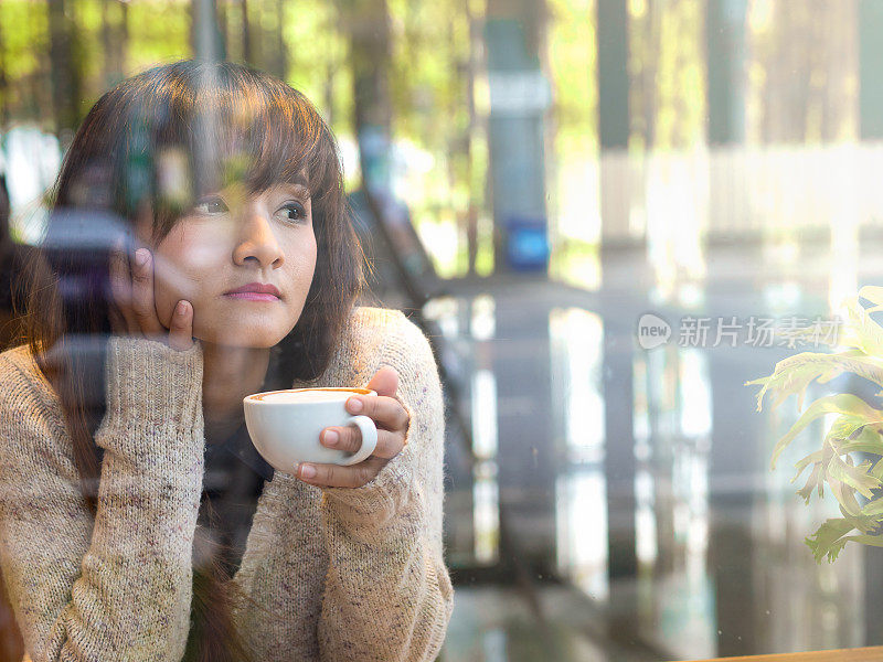 孤独的亚洲女人喝热咖啡在玻璃屋咖啡馆看着窗外等待某人
