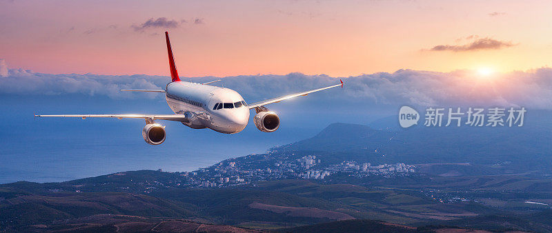 乘飞机。风景与白色的飞机是飞行在橙色的天空与云的山，海在五颜六色的日落。客机正在降落。商用飞机。私人飞机。旅行