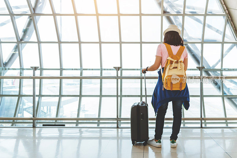 亚洲年轻女子与手提箱和黄色背包在机场窗口等待航班。