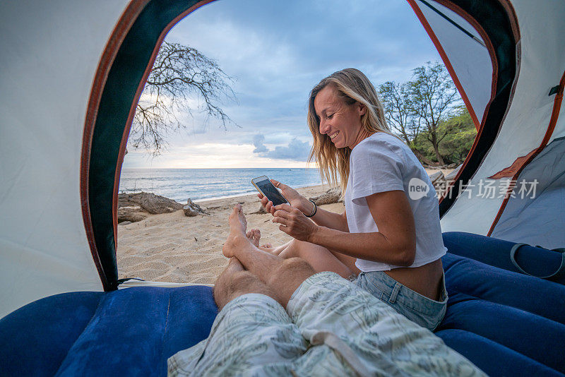 年轻夫妇在帐篷里用手机，两人在沙滩上露营