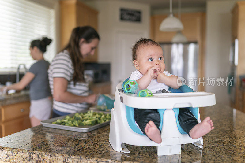 小女孩坐在厨房柜台上的婴儿椅上，其他人在做午饭