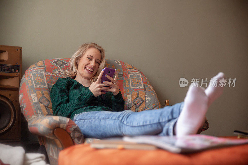 一个年轻的女人真的很快乐，她坐在沙发上，一边笑，一边用她的智能手机发短信。