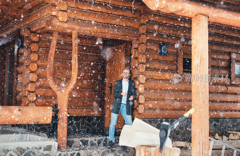 一个男人呆在乡下房子的门廊上享受降雪。木质树桩与斧头在下雪的院子里，不聚焦与沉重的雪花。冬季乡村假日概念形象