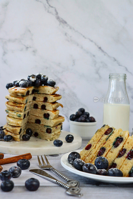图片堆叠的美式蓝莓煎饼与新鲜浆果，在大理石蛋糕架和旁边的盘子煎饼片，碗蜂蜜和木制蜂蜜勺，牛奶瓶，周围的新鲜蓝莓，大理石效果的背景，聚焦前景