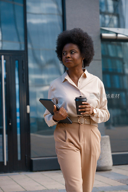 一位年轻的深色皮肤的女商人，穿着经典的米色套装，留着非洲式的头发，端着一杯茶和一台数码平板电脑走在街上