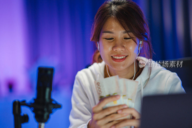年轻的亚洲妇女拿着钱银行票据和录制播客在她的笔记本电脑在家庭工作室在晚上。