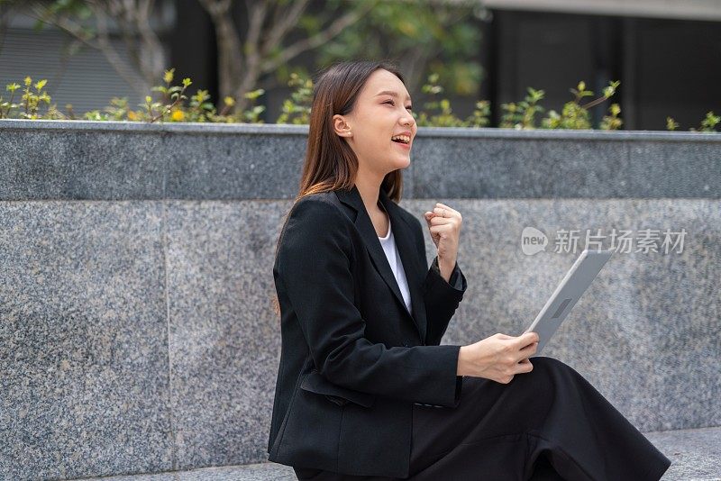 年轻美丽的亚洲女性办公室行政人员显示yes获胜的姿态，在她的办公大楼外，在她的平板电脑上与她的队友聊天