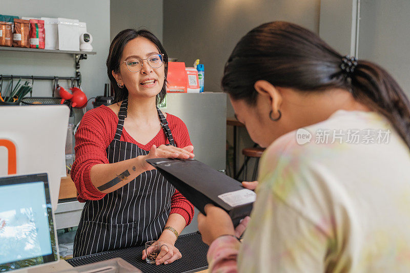 年轻的亚洲女性咖啡师或咖啡店老板在咖啡馆的吧台讨论点饮料菜单并向她的客户推荐咖啡豆