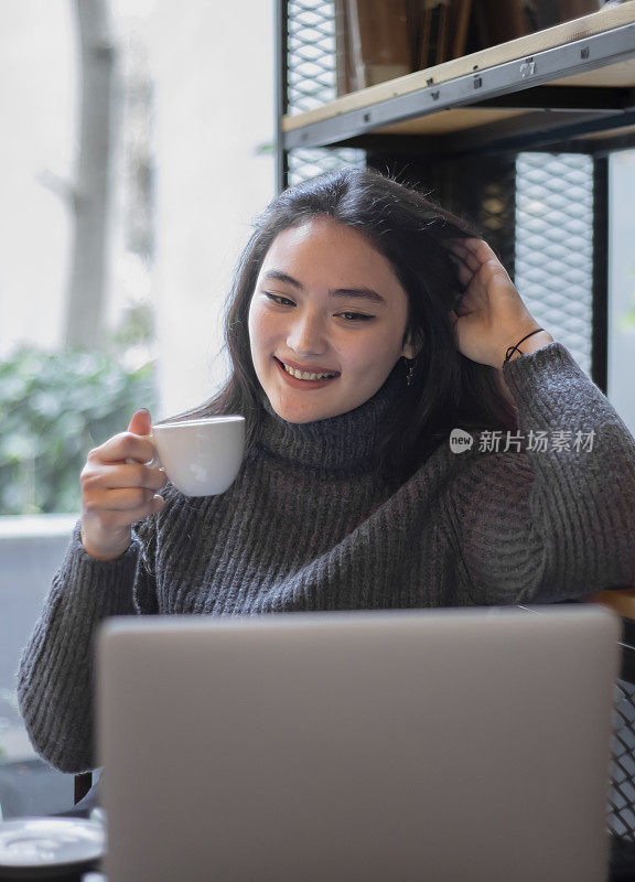 一名年轻的亚洲女子在咖啡馆边喝咖啡边摆弄电脑