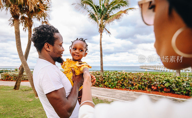 非洲裔美国父亲和母亲在热带地区与可爱的蹒跚学步的女儿玩耍，家庭度假和现代男子气概