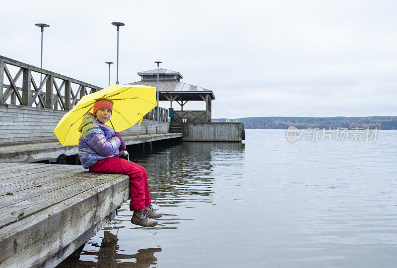 一个7岁的白人女孩打着明黄色的雨伞，坐在湖边的木码头上做梦。秋天的心情。