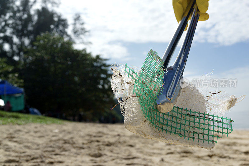 一名志愿者用钳子清理海滩上的塑料碎片和垃圾的特写