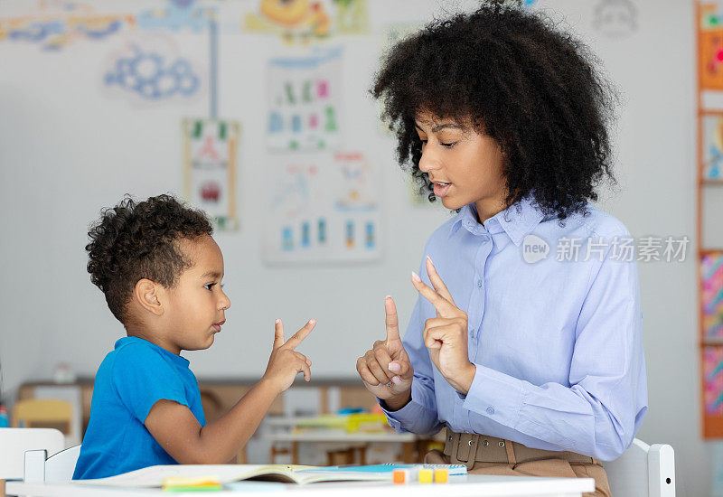小黑人男孩在女老师的帮助下学习用手指数数，坐在教室里的课桌前