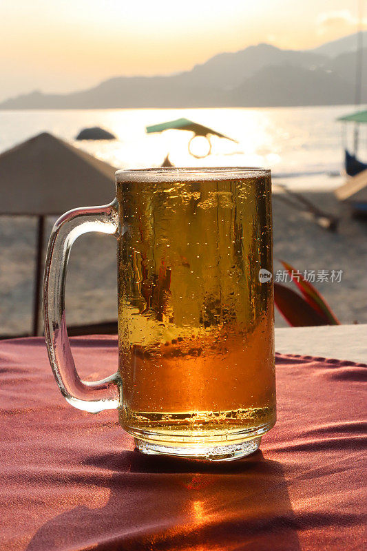 啤酒在玻璃大酒杯的形象覆盖在凝结与起泡的气泡形成泡沫的啤酒顶部的泡沫，马克杯品脱玻璃杯在海滩酒吧户外桌子与桌布，遮阳伞在沙滩上日落，停泊的渔船，重点在前景