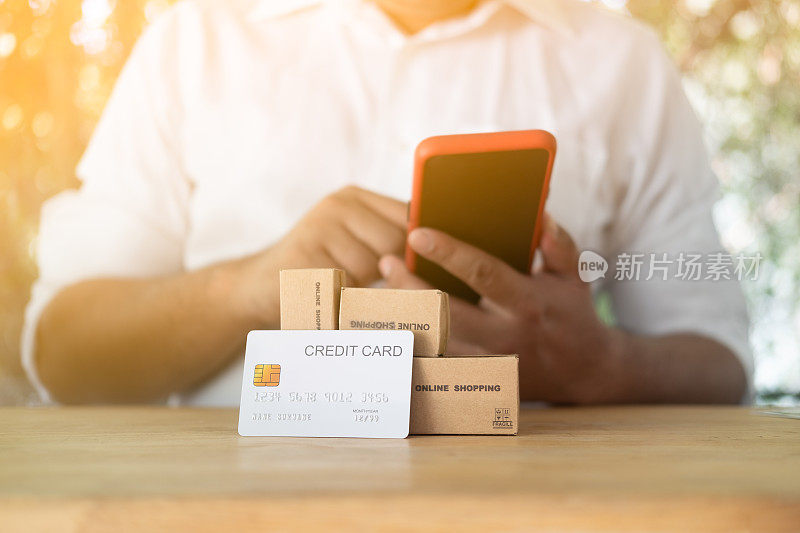 人们用手用手机进行在线支付，迷你棕色盒子和白色信用卡放在前面。通过手机银行应用在线支付的概念，网上购物的概念。