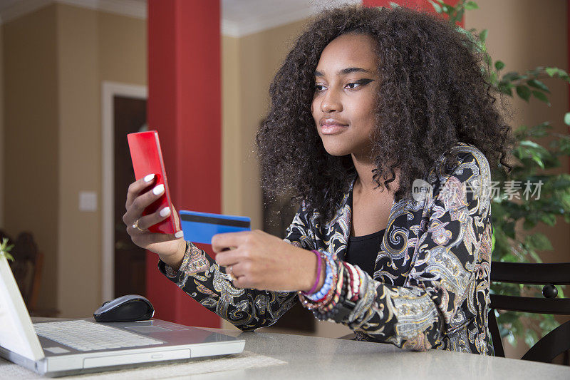 可爱的非裔美国青少年在网上购物。她坐在厨房吧台旁。笔记本电脑、智能手机和信用卡。