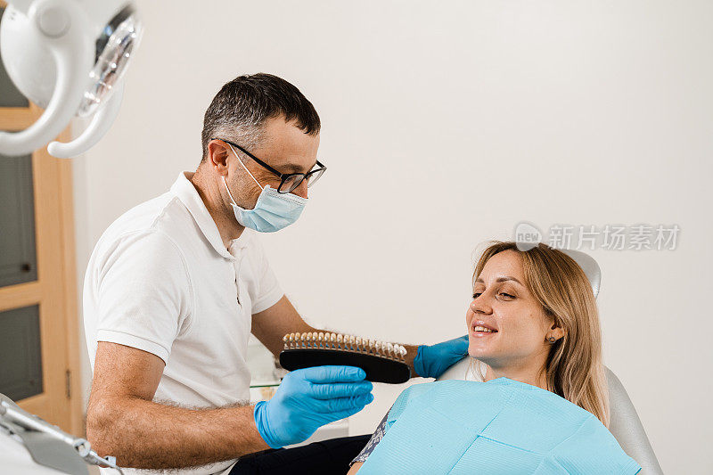 妇女看着贴面或种植牙配色样本在医生手中。牙医。牙科医生在牙科诊所为女性病人示范牙齿美白的颜色深浅。