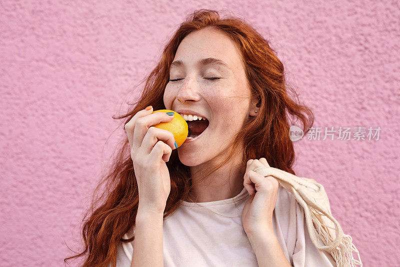 红头发的女人在粉红色的城墙上咬着美味的橙子