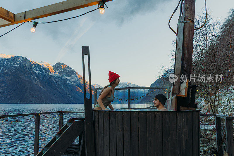 在挪威的冬天，一名女子和一名男子在峡湾边的热水浴缸里放松，欣赏风景优美的山景