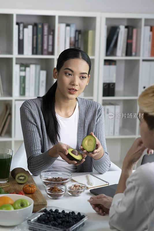 亚洲女性专业营养师与她的客户进行咨询，桌子上摆满了各种水果、坚果、蔬菜和膳食补充剂