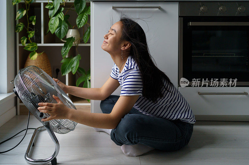 开朗积极的亚洲女孩享受冷风从电扇坐在地板上的房子的厨房