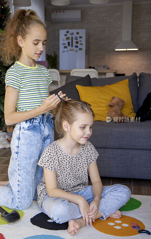 两个小姐妹坐在客厅的地板上，做着美丽而有趣的发型。大一点的给小一点的戴上发带。早晨的时间和友谊的概念